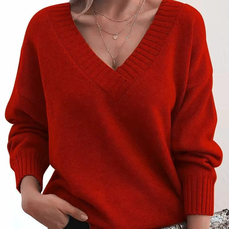 Une femme porte un pull à col V éclatant, à manches longues et à la coupe ajustée. Elle arbore plusieurs colliers en or délicats et a une main glissée dans son pantalon, ce qui lui confère une allure élégante et moderne.