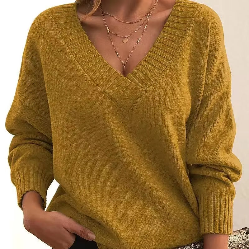 Femme portant un pull moutarde à col en V, avec des manches longues et une coupe slim. Elle porte plusieurs colliers fins en or et une main est glissée dans son pantalon, affichant un look moderne et élégant.