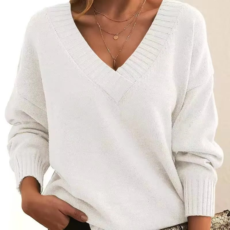 Femme portant un pull blanc à col en V, avec des manches longues et une coupe slim. Elle porte plusieurs colliers fins en or, et une main est glissée dans son pantalon, affichant un style moderne et élégant.