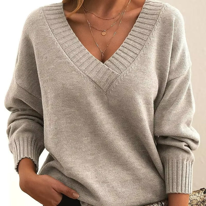 Une femme porte un pull beige à col en V, à manches longues et à la coupe ajustée. Elle est parée de plusieurs délicats colliers en or et une main est glissée dans son pantalon, créant une allure à la fois élégante et moderne.