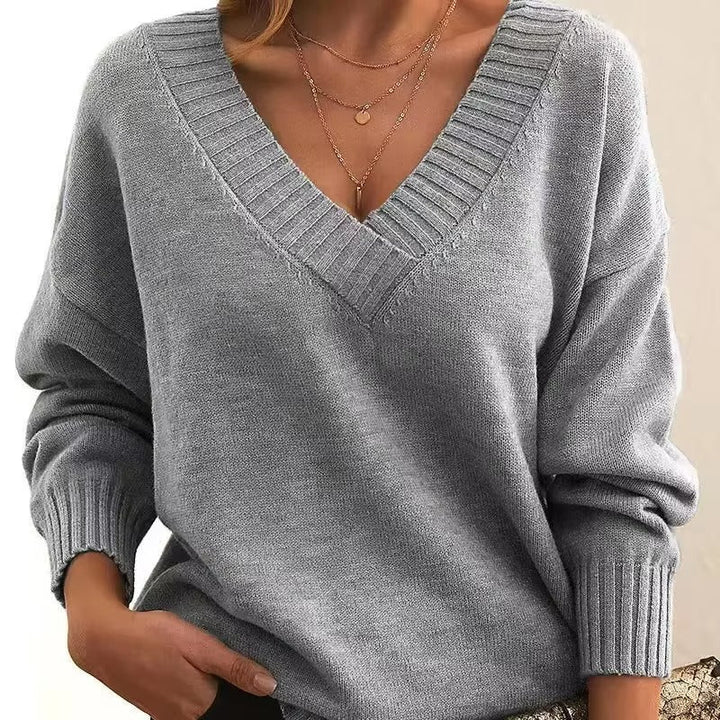 Femme portant un pull gris clair à col en V, avec des manches longues et une coupe slim. Elle porte plusieurs colliers délicats en or, et une de ses mains est glissée dans son pantalon, affichant un style moderne et épuré.