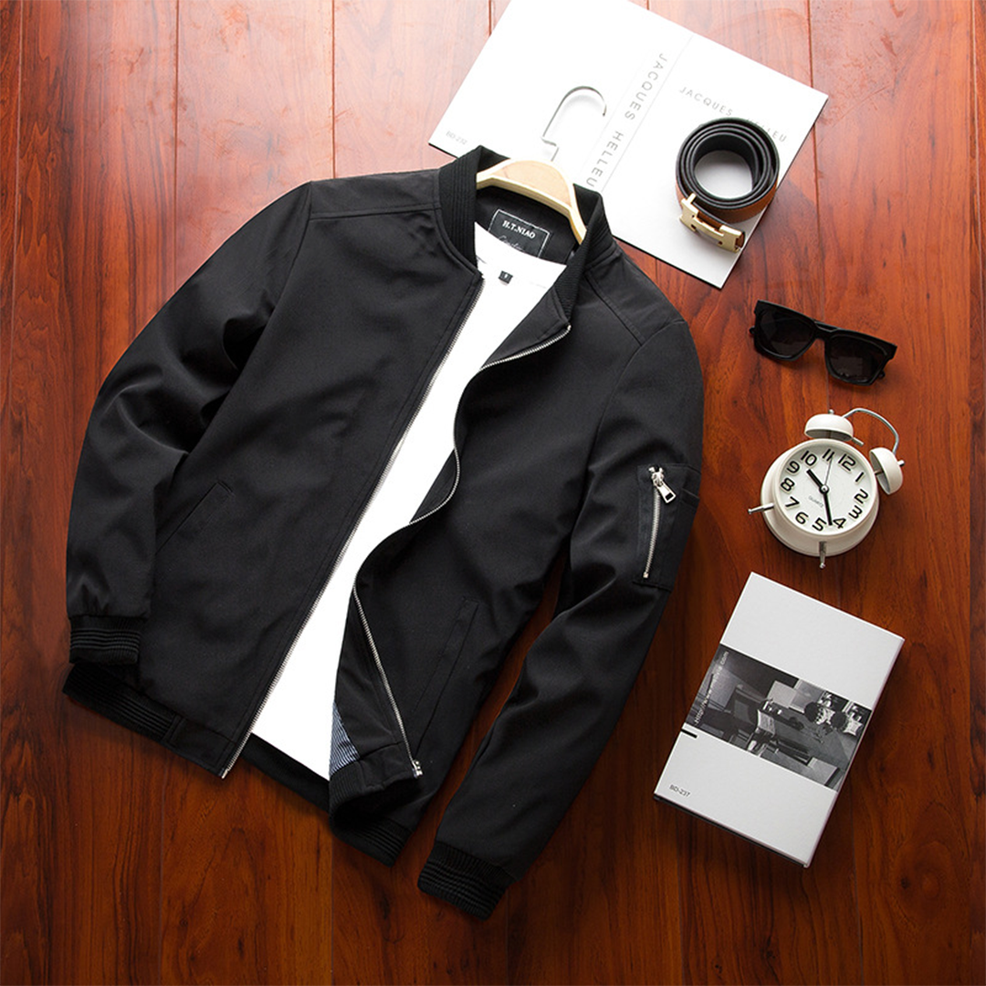 Veste bomber noire légère avec fermeture zippée, col et poignets côtelés, poches latérales et poche zippée sur la manche, photographiée à plat sur une table en bois avec accessoires.