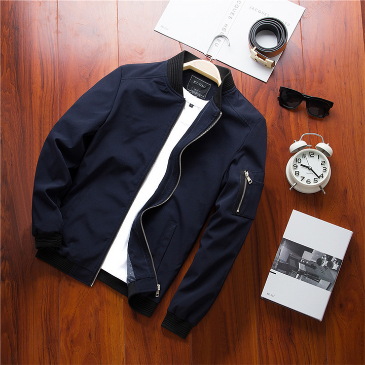 Veste bomber bleu marine avec col et poignets côtelés noirs, fermeture zippée, poches latérales et poche zippée sur la manche, présentée à plat sur table en bois avec accessoires.