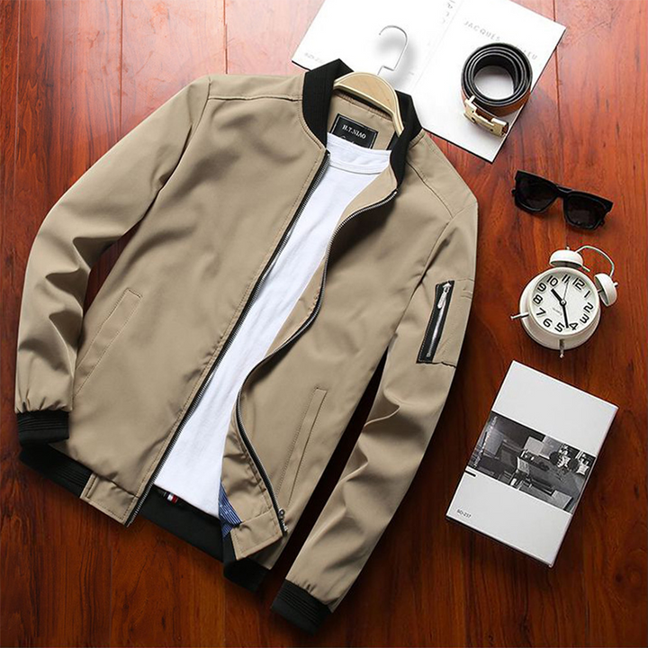 Veste bomber beige/kaki clair avec col et poignets côtelés noirs, fermeture zippée, poches latérales et poche zippée sur la manche, présentée à plat sur table en bois avec accessoires.