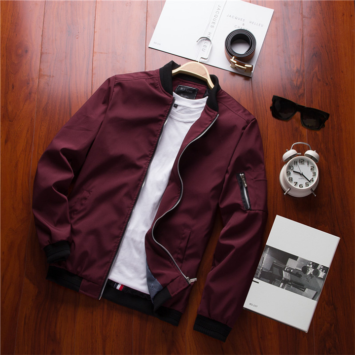 Veste bomber bordeaux avec col et poignets côtelés noirs, fermeture zippée, poches latérales et poche zippée sur la manche, présentée à plat sur table en bois avec accessoires.