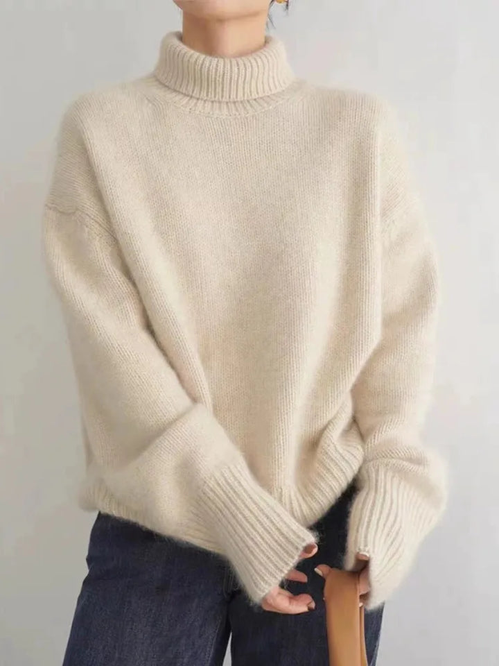 Pull d’hiver pour femme en maille douce de couleur écru, avec col roulé épais, coupe ample et manches longues aux poignets côtelés, porté avec un jean pour un look minimaliste et chaleureux.