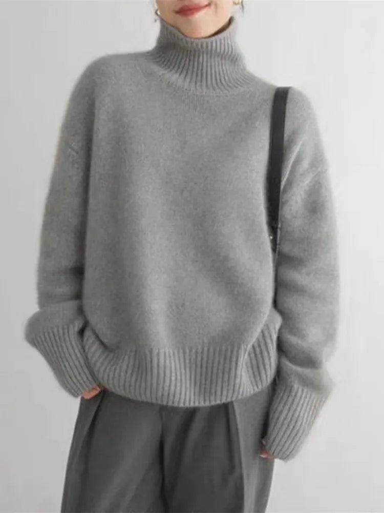 Pull d’hiver pour femme en maille douce de couleur gris clair, avec col roulé épais, coupe ample et manches longues aux poignets côtelés, porté avec un pantalon fluide pour un style élégant et confortable.