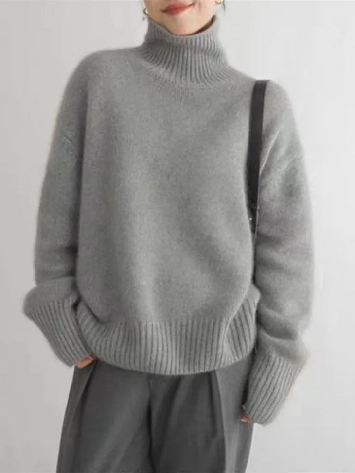Pull d’hiver pour femme en maille douce de couleur gris clair, avec col roulé épais, coupe ample et manches longues aux poignets côtelés, porté avec un pantalon fluide pour un style élégant et confortable.
