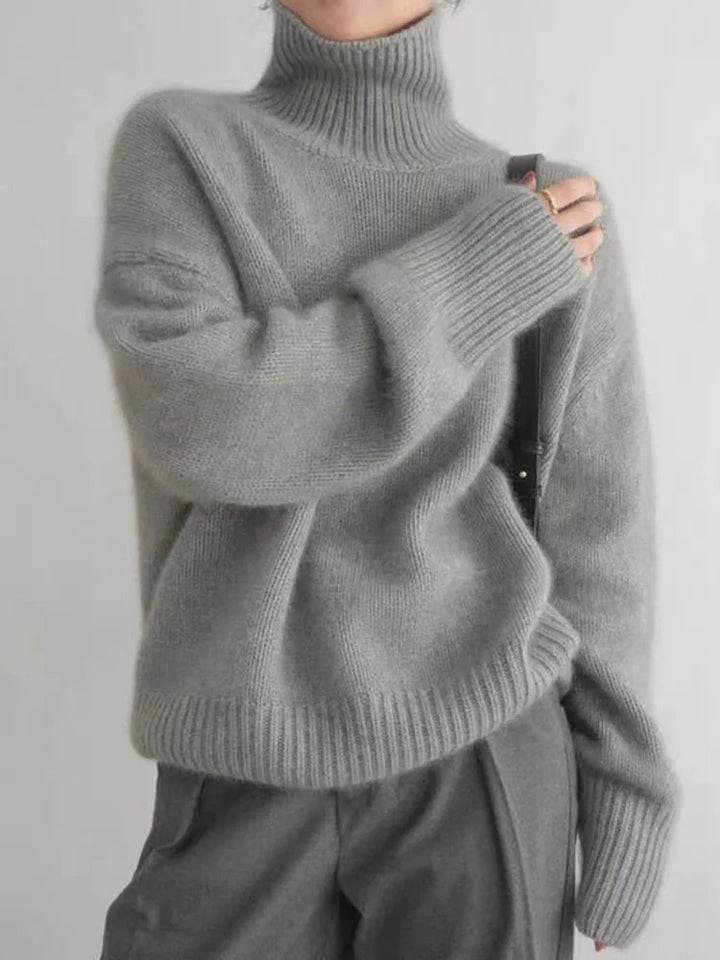 Pull d’hiver pour femme en maille douce de couleur gris moyen, avec col roulé montant, coupe ample et manches longues aux poignets côtelés, porté avec un pantalon assorti pour un look moderne et chaleureux.