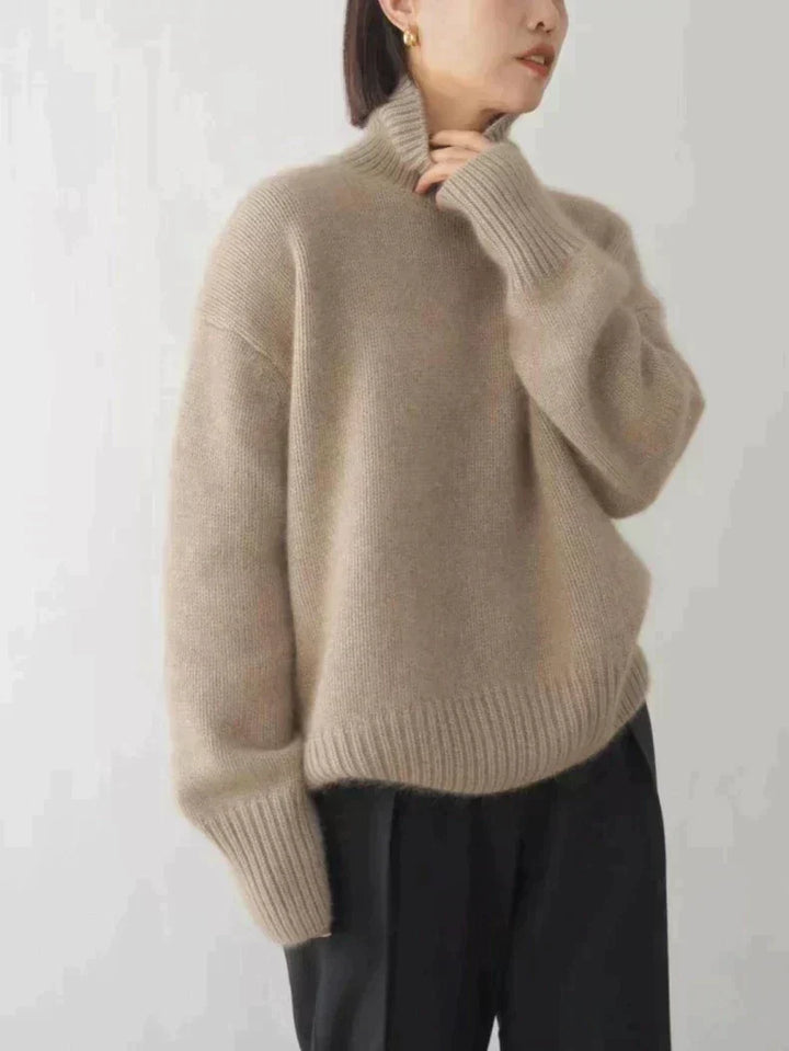 Pull d’hiver pour femme en maille douce de couleur beige sable, avec col roulé épais, coupe ample et manches longues aux poignets côtelés, porté avec un pantalon noir pour un style élégant et confortable.