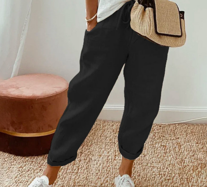 Pantalon ample pour femme noir en tissu léger, coupe large et confortable avec taille élastiquée et cordon de serrage, porté avec des baskets blanches et un sac en paille pour un look décontracté et moderne.