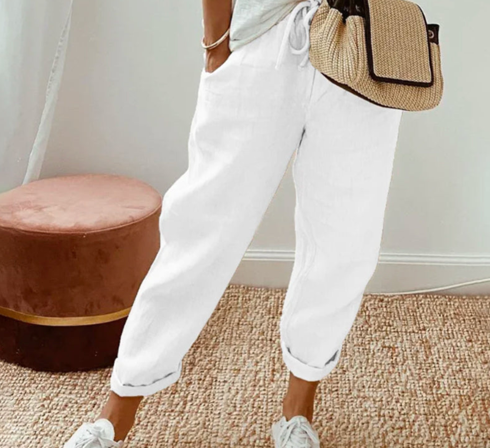 Pantalon ample pour femme blanc en tissu léger, coupe large et confortable avec taille élastiquée et cordon de serrage, porté avec des baskets blanches et un sac en paille pour un look frais, décontracté et moderne.