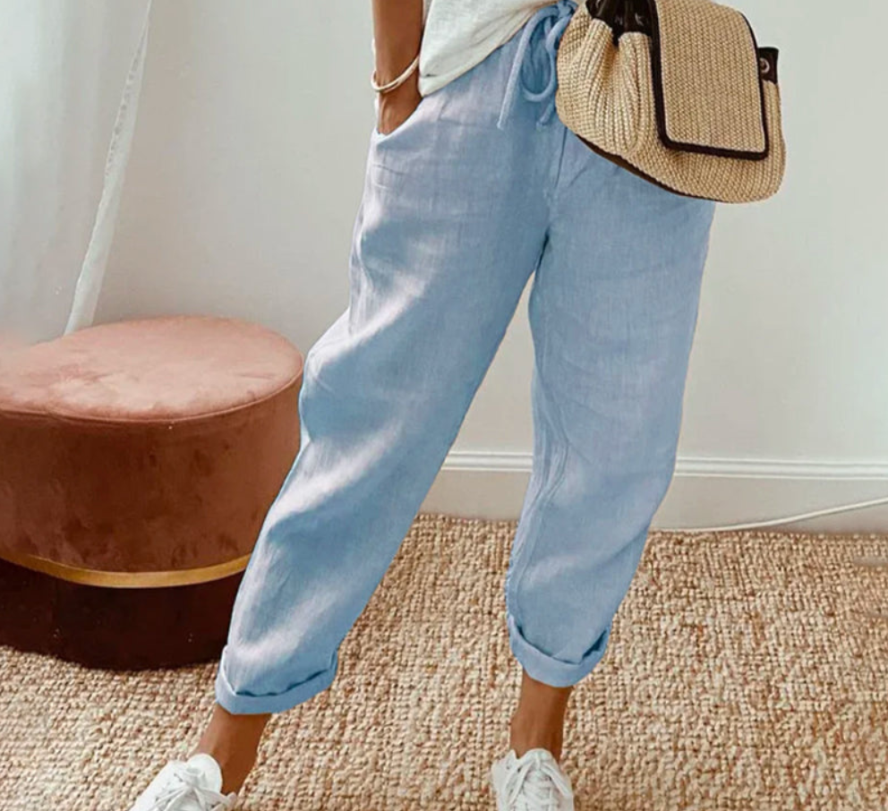 Pantalon ample pour femme bleu clair en tissu léger, coupe large et confortable avec taille élastiquée et cordon de serrage, porté de manière décontractée avec des baskets blanches et un sac en paille pour un style estival et moderne.