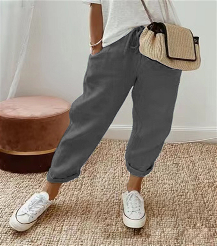 Pantalon ample pour femme gris foncé en tissu léger, coupe large et confortable avec taille élastiquée et cordon de serrage, porté avec un haut clair, des baskets blanches et un sac en paille pour un style décontracté et moderne.