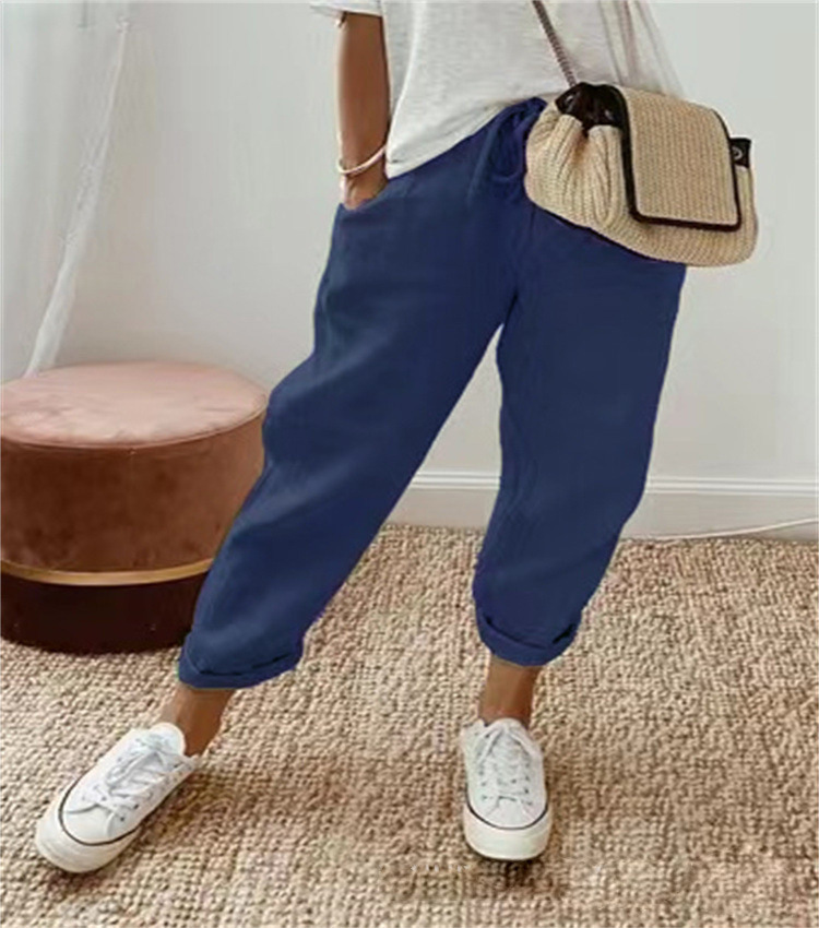 Pantalon ample pour femme bleu foncé en tissu léger, coupe large et confortable avec taille élastiquée et cordon de serrage, porté avec un haut clair, des baskets blanches et un sac en paille pour un look décontracté et moderne.