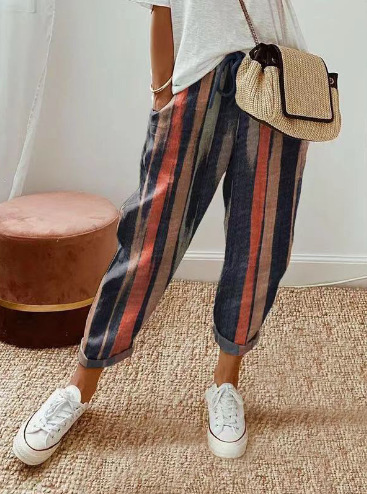 Pantalon ample pour femme en tissu léger à rayures verticales bleu foncé, terracotta et beige, coupe large et confortable avec taille élastiquée et cordon de serrage, porté avec des baskets blanches et un sac en paille pour un style décontracté et tendance.