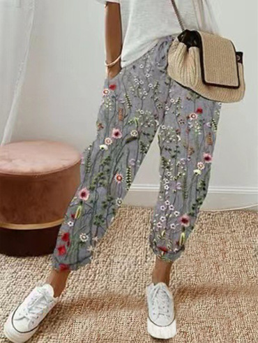 Pantalon ample pour femme en tissu léger gris à motif floral multicolore, coupe large et confortable avec taille élastiquée et cordon de serrage, porté avec un haut clair, des baskets blanches et un sac en paille pour un look décontracté et féminin.