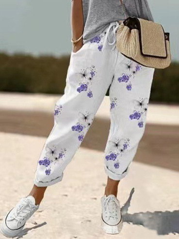 Pantalon ample pour femme blanc en tissu léger à motif floral violet et noir, coupe large et confortable avec taille élastiquée et cordon de serrage, porté avec un haut gris, des baskets blanches et un sac en paille pour un style estival et décontracté.