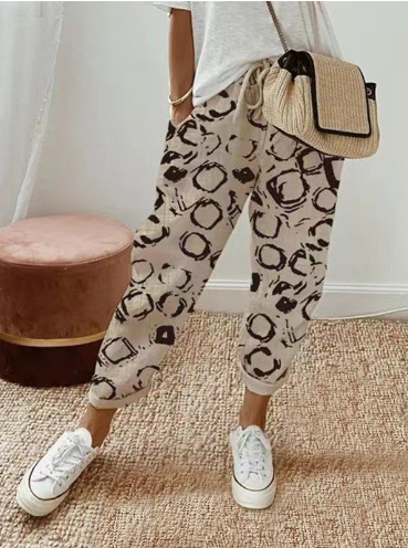 Pantalon ample pour femme en tissu léger beige à motifs abstraits marron, coupe large et confortable avec taille élastiquée et cordon de serrage, porté avec un haut clair, des baskets blanches et un sac en paille pour un look décontracté et tendance.