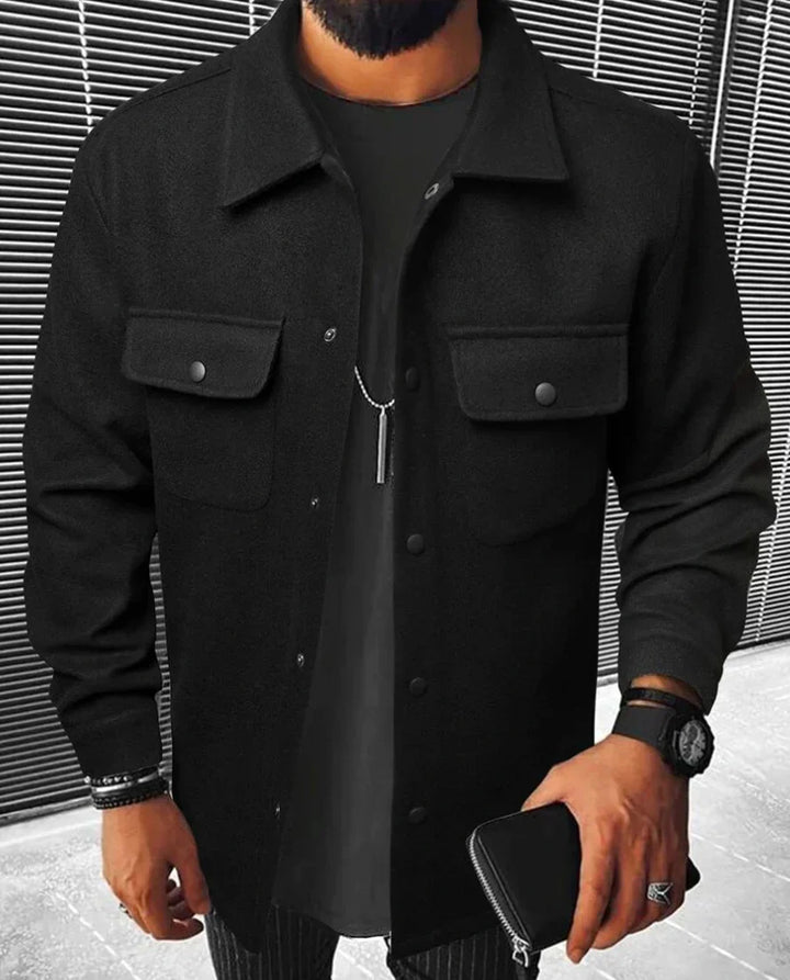 Homme portant une chemise noire à deux poches sur la poitrine, avec un t-shirt noir en dessous et un accessoire de montre, tenant un portefeuille en cuir, dans un décor urbain moderne.