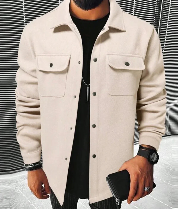 Homme portant une chemise beige à boutons noirs et deux poches sur la poitrine, un t-shirt noir en dessous, avec une montre et un portefeuille en cuir dans la main, posé dans un environnement moderne.