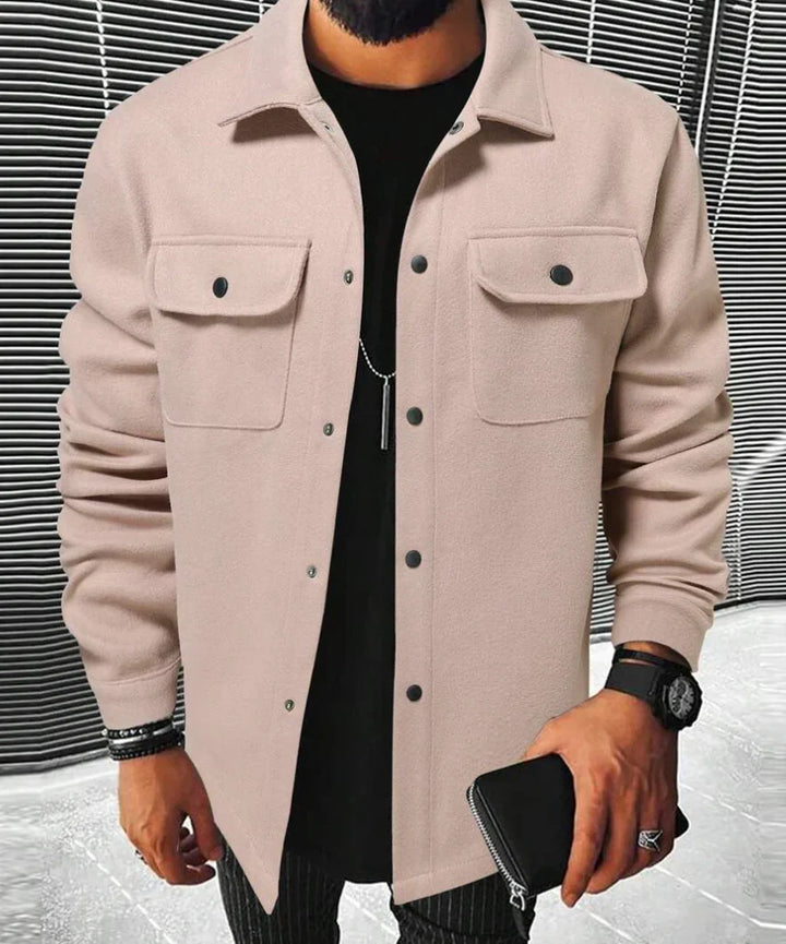 Homme portant une chemise beige à deux poches sur la poitrine, un t-shirt noir en dessous, une montre et un portefeuille en cuir, posé dans un environnement moderne.