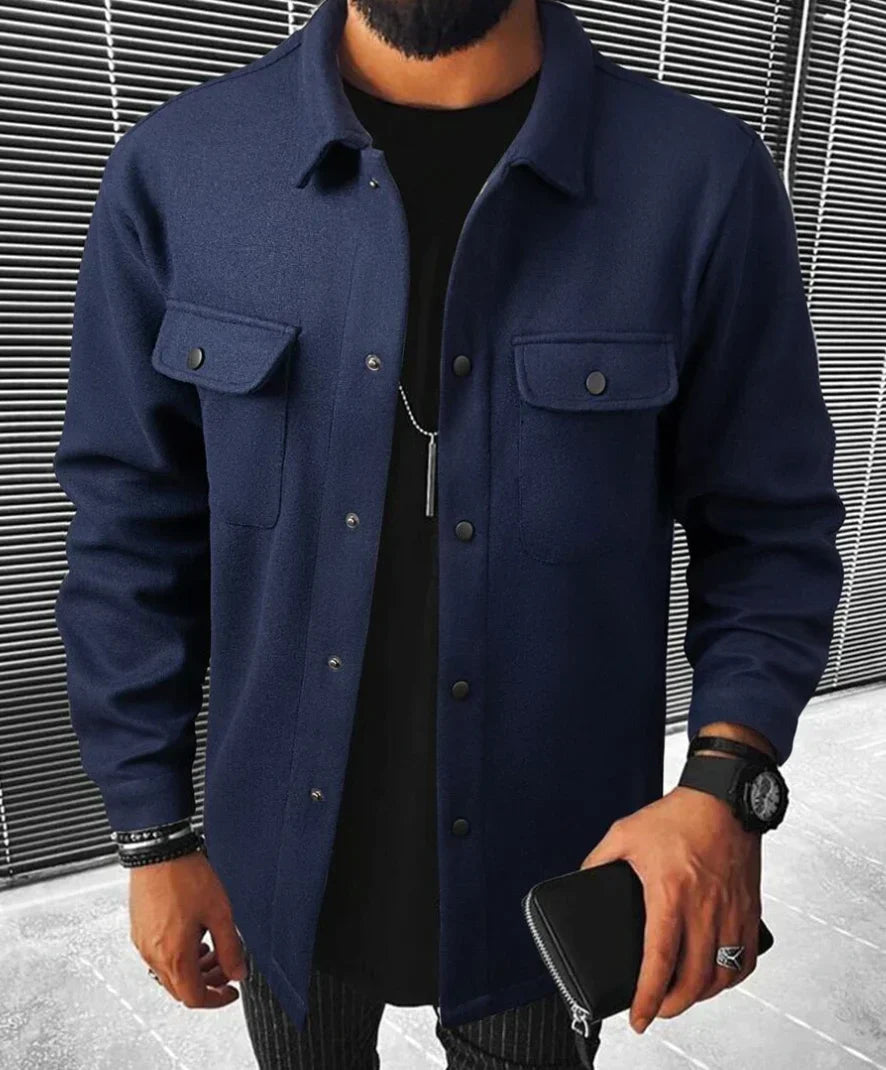 "Homme portant une chemise bleu marine avec des boutons noirs et deux poches sur la poitrine, avec un t-shirt noir en dessous. Il tient un portefeuille en cuir et porte une montre, dans un cadre moderne.