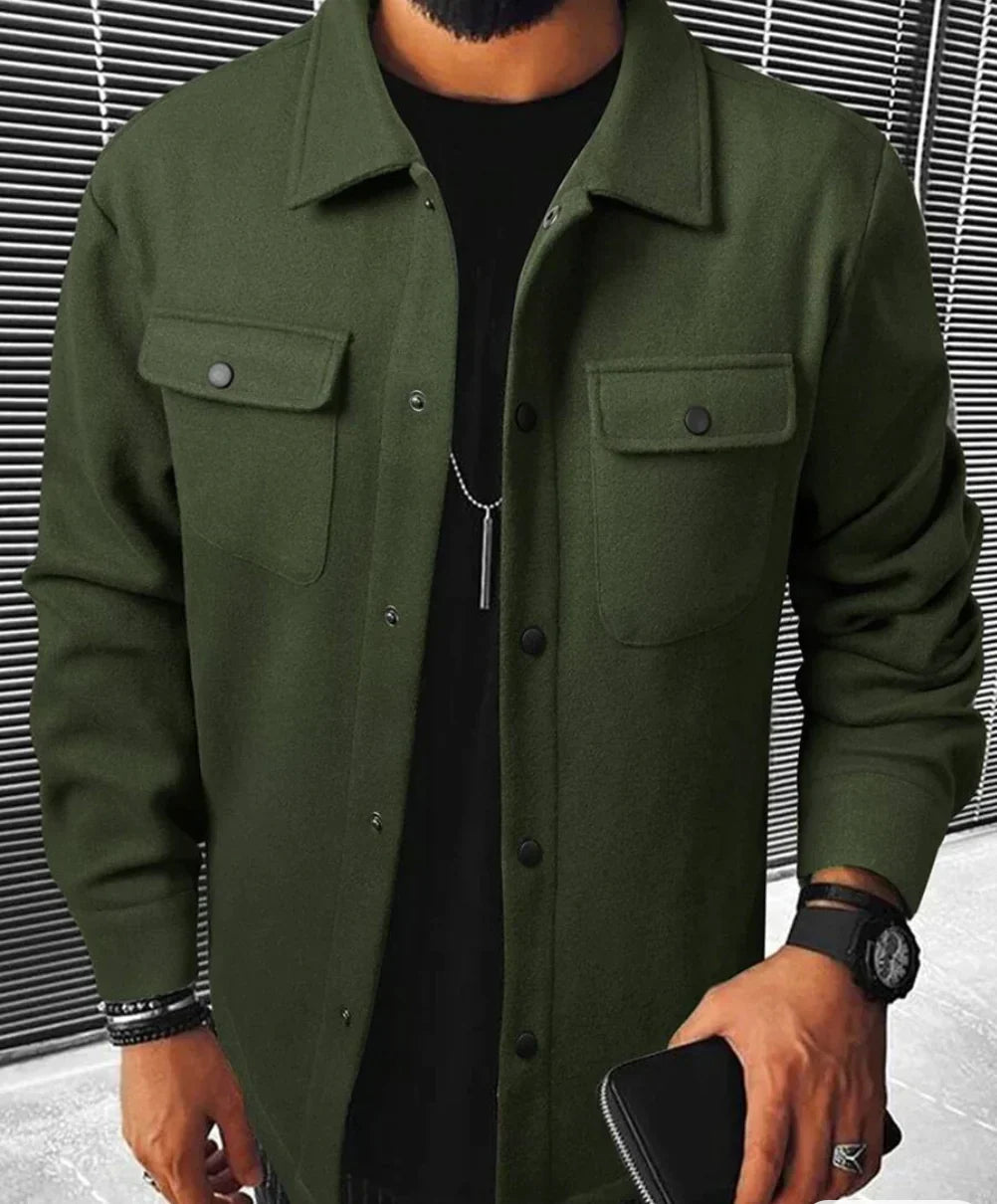 Homme portant une chemise verte à boutons noirs avec des poches sur la poitrine et un style sobre et moderne. Il est habillé d’un t-shirt noir en dessous et porte des accessoires tels qu'une montre et des bracelets. Le fond minimaliste avec des lignes grises ajoute à l’élégance discrète de la tenue. Une combinaison parfaite de confort et de style décontracté pour un look raffiné.