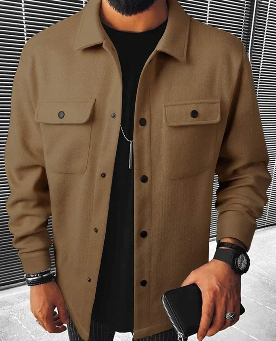Homme portant une chemise marron à boutons noirs, avec des poches sur la poitrine et un design simple et élégant. Il est habillé avec un t-shirt noir en dessous et porte une montre et des bracelets. Le fond montre des lignes grises modernes, créant un contraste avec l’aspect classique de la chemise. Cette tenue allie confort et style pour un look décontracté et sophistiqué.