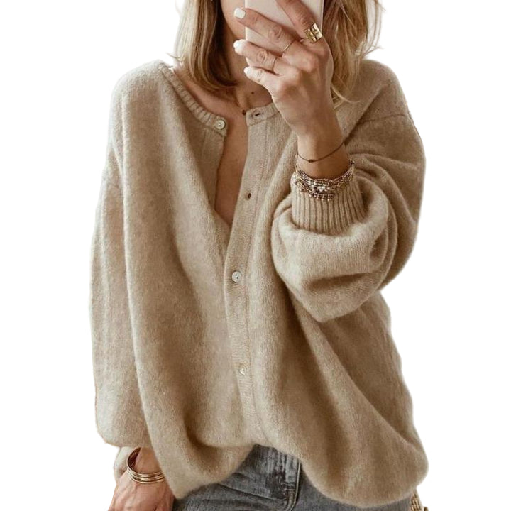 Une femme portant un long cardigan marron en maille douce, associé à un jean taille haute, pose dans une chambre moderne tout en prenant une photo avec son téléphone.