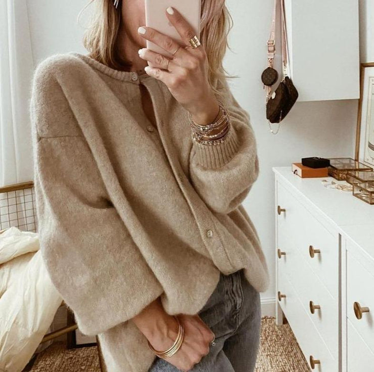 Une femme portant un cardigan en maille marron clair, légèrement ouvert sur un haut assorti, associé à un jean taille haute, pose dans une chambre moderne tout en prenant une photo avec son téléphone.