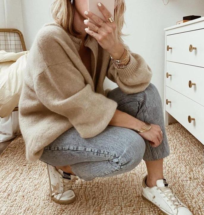 Une femme accroupie, vêtue d'un cardigan en maille marron clair, d'un jean clair et de baskets blanches, pose dans une chambre de style moderne tout en prenant une photo avec son téléphone.