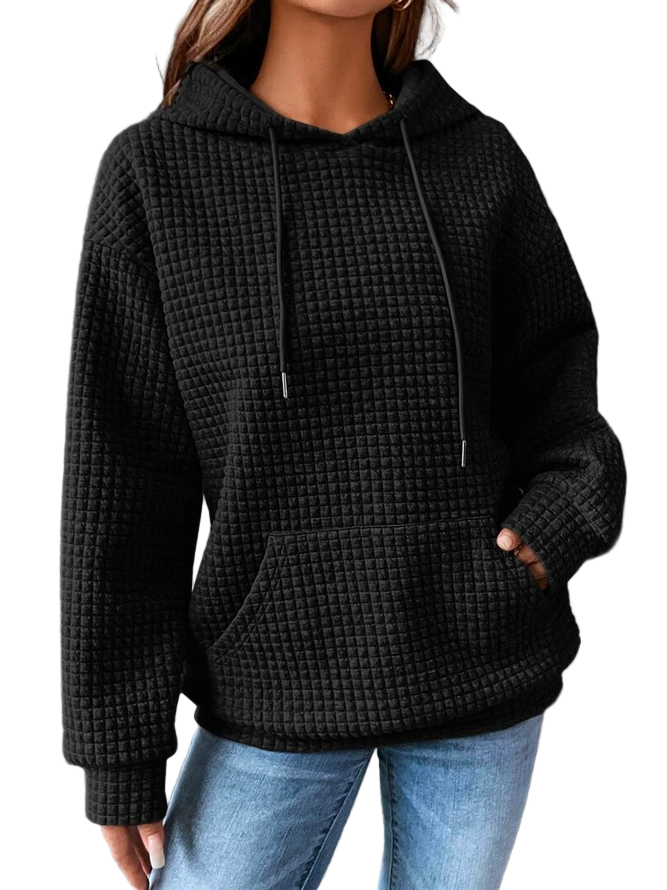 Femme portant un sweat à capuche noir à texture gaufrée, doté d’une grande poche frontale et de cordons ajustables. Le tissu épais et doux offre une allure cosy et moderne, idéale pour affronter les journées froides d’hiver. Associé à un jean clair, ce vêtement crée un look décontracté et élégant, parfait pour un style urbain confortable.