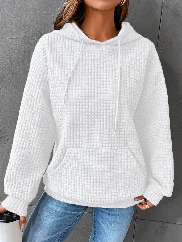 Femme portant un sweat à capuche blanc texturé, au tissu chaud et moelleux. Le vêtement présente une coupe ample avec des cordons de serrage et une grande poche avant, parfait pour un style décontracté et confortable durant la saison hivernale. Associé à un jean bleu, cet ensemble offre un look cosy et moderne, idéal pour les journées froides.
