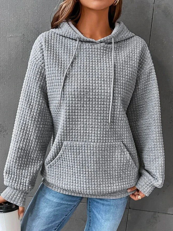 Une femme porte un sweat à capuche gris gaufré, doté d'une grande poche avant et de cordons de serrage ajustables. Son tissu épais et doux offre chaleur et confort, idéal pour les mois les plus froids. Associé à un jean bleu, ce look décontracté et moderne est parfait pour les journées d'hiver.