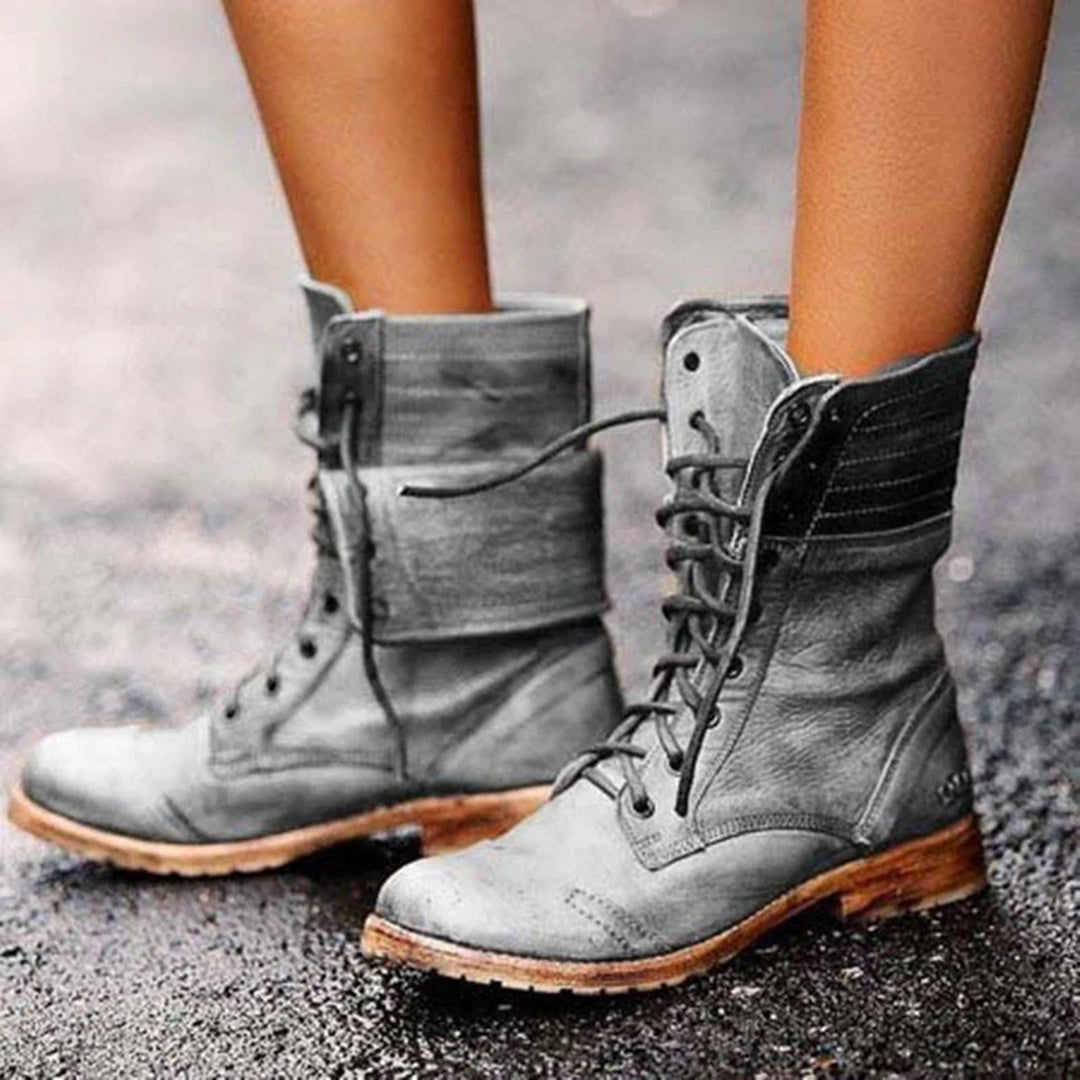 Une paire de bottines montantes en cuir gris clair avec lacets, au style vintage et semelle en bois naturel. Les chaussures présentent un effet vieilli élégant, idéal pour un look décontracté et robuste. Elles sont portées en extérieur, soulignant leur aspect polyvalent et tendance pour la saison automne-hiver.
