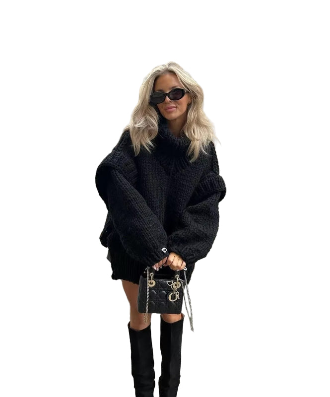 Femme posant dans une rue élégante, portant un pull noir oversize en maille épaisse avec col montant. Elle accessoirise sa tenue avec des lunettes de soleil, un sac à main noir structuré et des bottes hautes assorties. Son look d’hiver chic contraste avec l’architecture classique des immeubles en arrière-plan.