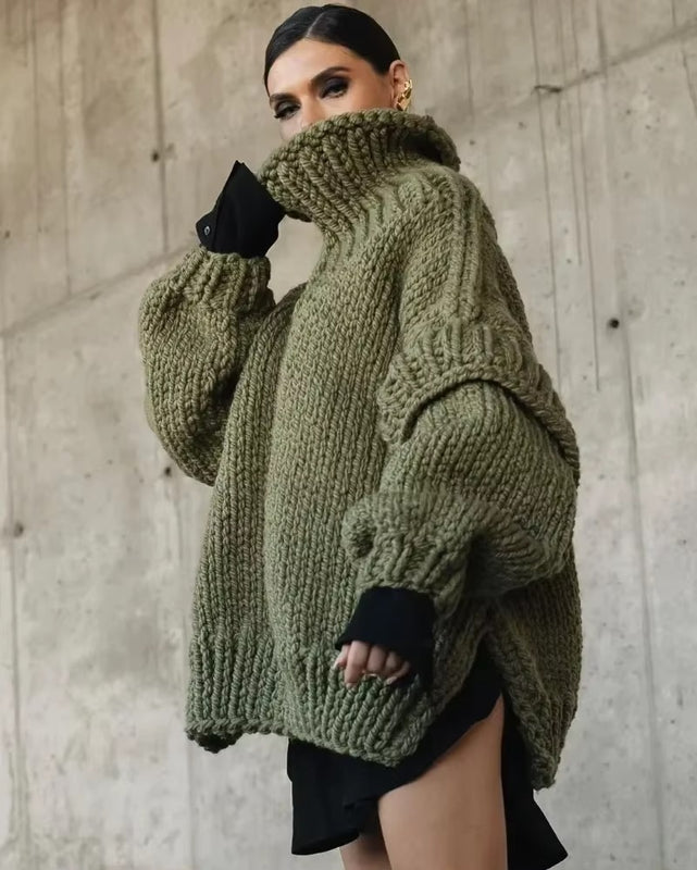 Une femme pose devant un mur en béton, vêtue d'un gros pull vert à col roulé volumineux. Les manches longues et amples accentuent l'effet oversize, tandis que la tenue noire en dessous apporte une touche moderne et contrastée. Son look est chic et audacieux, parfait pour l'hiver.