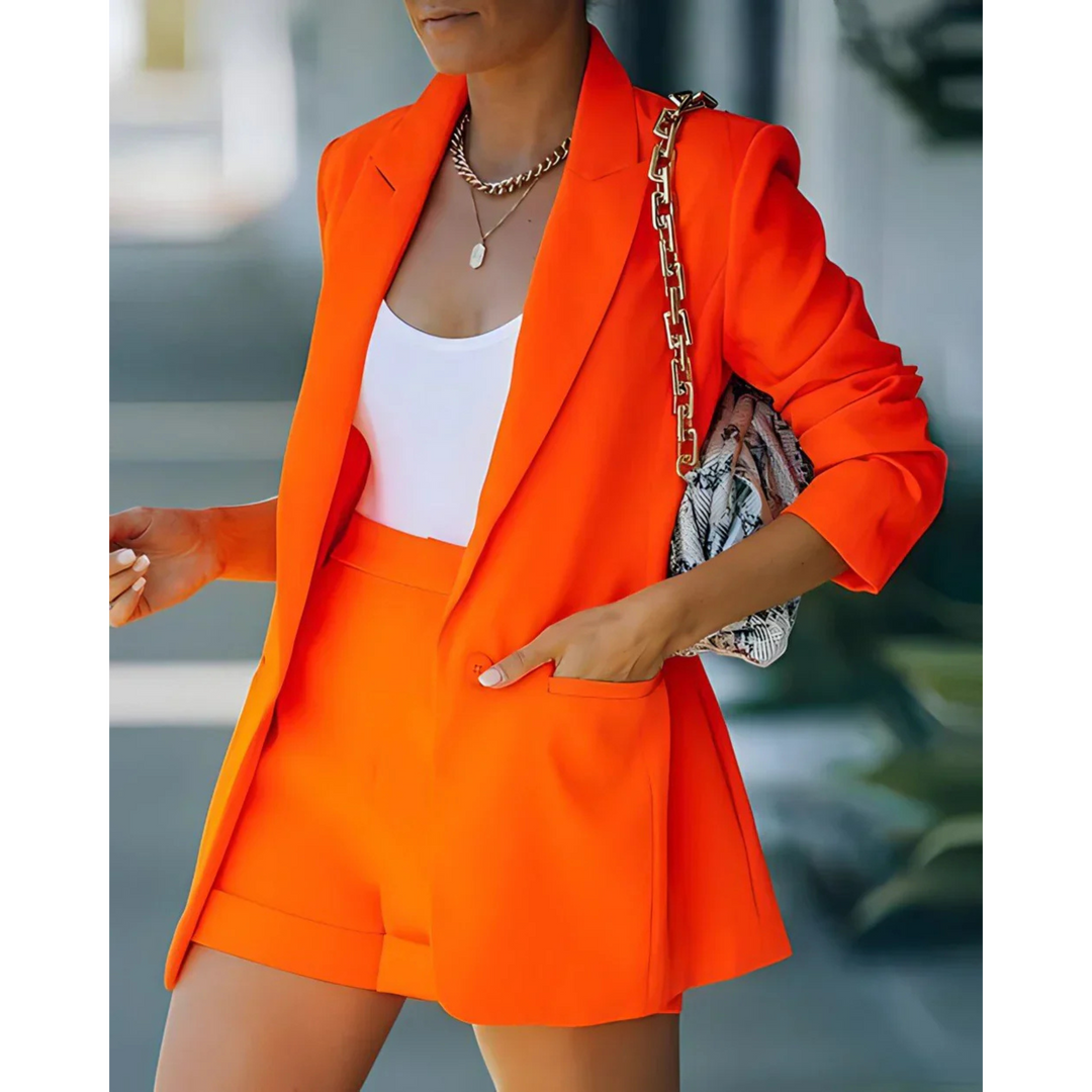 Blazer structuré pour femme de couleur orange vif, assorti à un short élégant et un top blanc. Le look est complété par un sac à main tendance, parfait pour un style moderne et dynamique, idéal pour les sorties estivales.