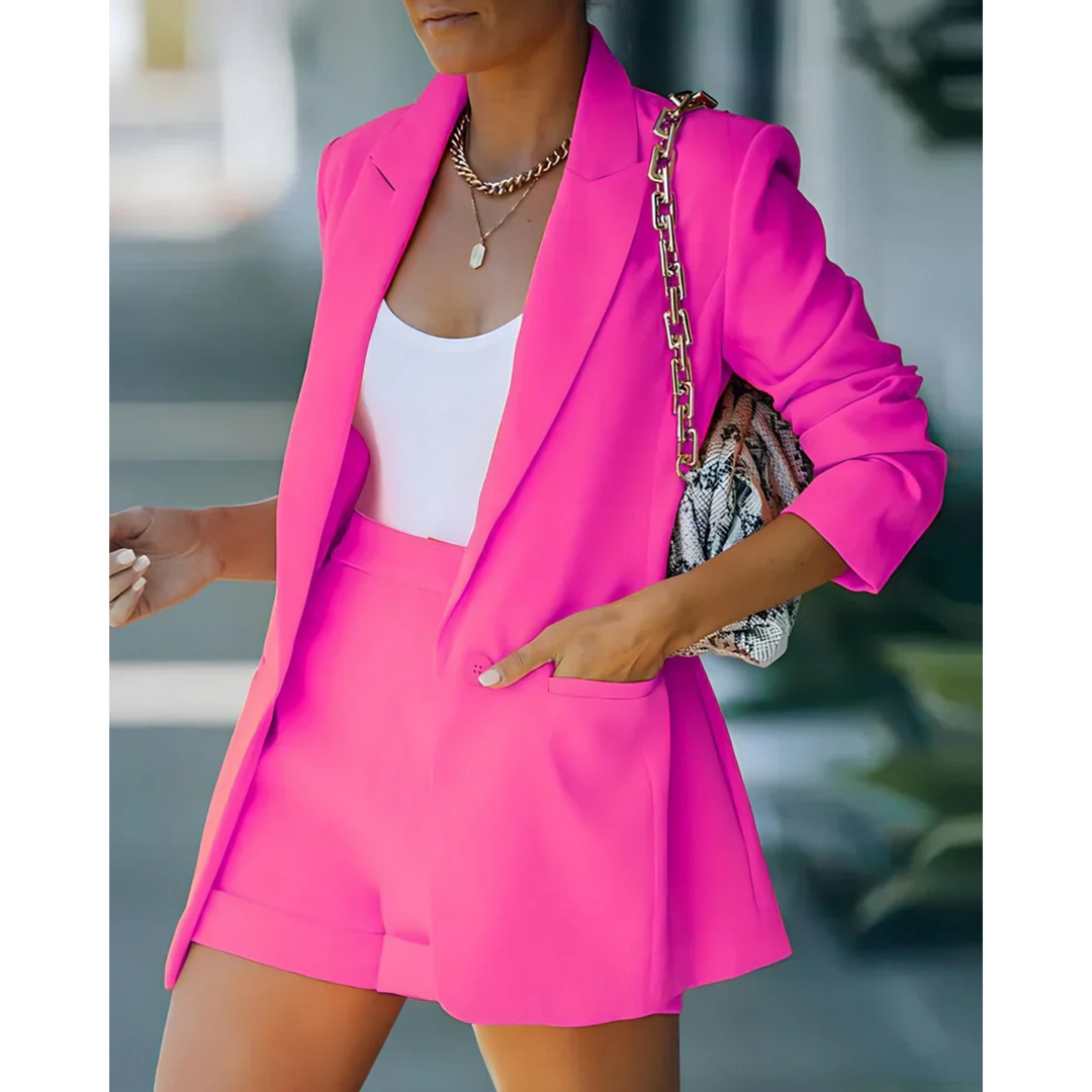 Blazer structuré pour femme de couleur fuchsia, accompagné d'un short assorti et d'un top blanc. Le look est complété par un sac à main à chaîne, parfait pour une tenue moderne, élégante et dynamique, idéale pour les occasions estivales.