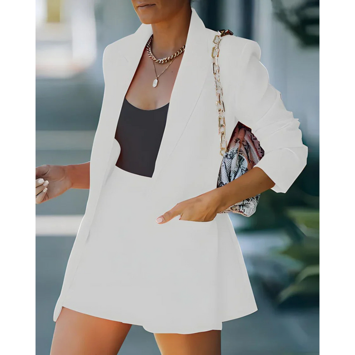 Blazer structuré pour femme de couleur blanche, accompagné d'un short assorti et d'un top noir. Le look est complété par un sac à main à chaîne, offrant une tenue moderne, élégante et parfaite pour un style estival chic.