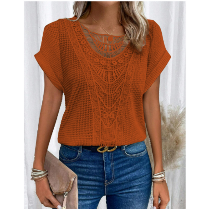 Femme portant le haut d’été Kester orange, coupe ample à manches courtes avec détail crochet ajouré au col, style tendance et léger, associé à un jean bleu et une pochette.