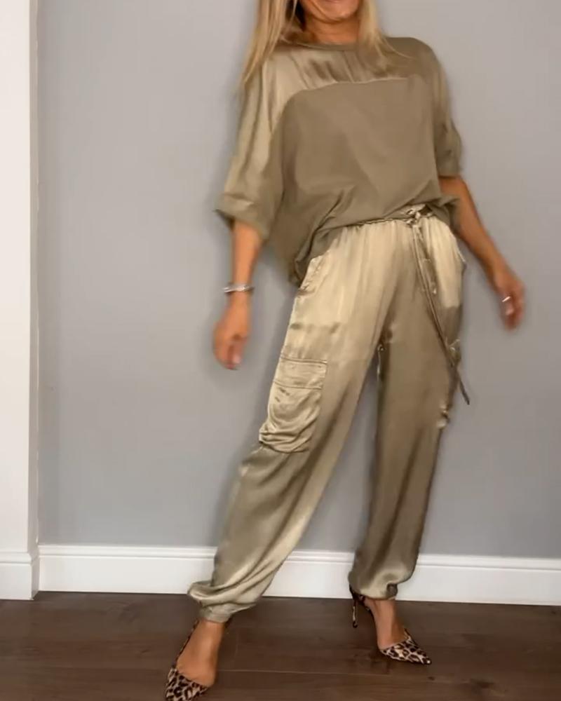 Une femme souriante portant un ensemble satiné beige avec un haut fluide et un pantalon avec des poches latérales. Elle pose en talons à imprimé léopard, créant un look élégant et décontracté.