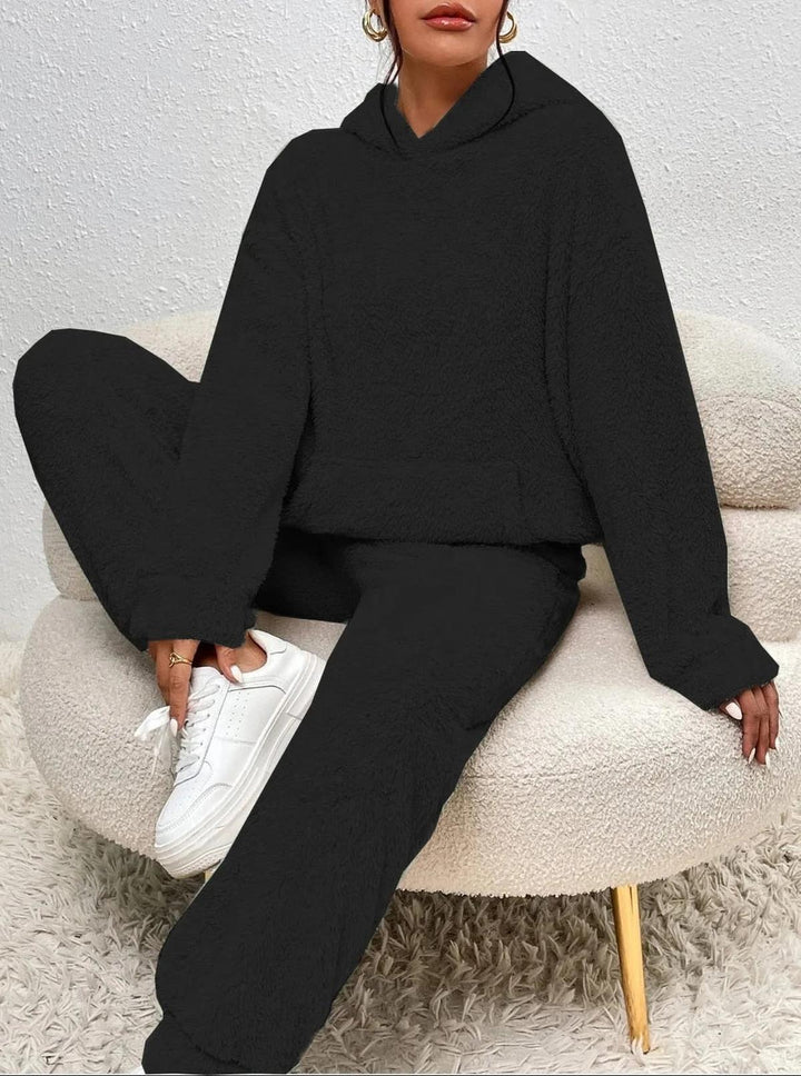 Ensemble loungewear pour femme en tissu teddy noir, composé d’un hoodie ample et d’un pantalon cosy, présenté en position assise sur un fauteuil doux, mettant en avant son confort moderne et son style décontracté.