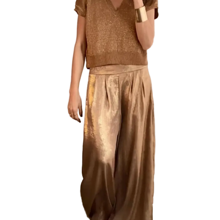 Femme portant un ensemble élégant composé d’un haut à manches courtes et d’un pantalon large taille haute, de couleur beige doré à l’aspect satiné, dans un intérieur raffiné au style chic et moderne.