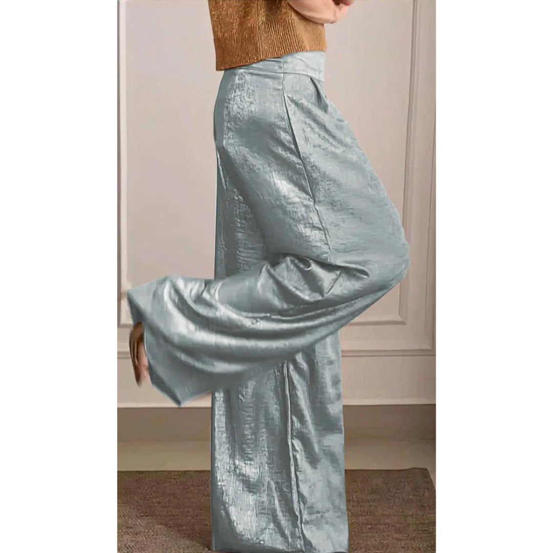 Femme portant un pantalon large taille haute de couleur bleu clair à l’aspect satiné, associé à un haut élégant, présenté de profil dans un intérieur chic et moderne.