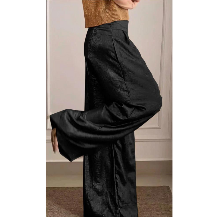 Femme portant un pantalon large taille haute de couleur noire à l’aspect satiné, associé à un haut élégant, présenté de profil dans un intérieur chic et moderne.
