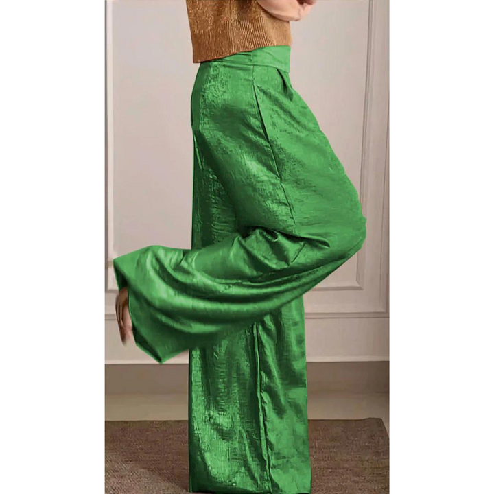 Femme portant un pantalon large taille haute de couleur verte à l’aspect satiné, associé à un haut élégant, présenté de profil dans un intérieur chic et moderne.