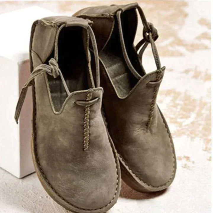Chaussures d’automne pour femme en cuir suédé gris taupe, avec bout arrondi, coutures apparentes et lien décoratif sur le côté, présentées sur un fond clair pour un style moderne et naturel.