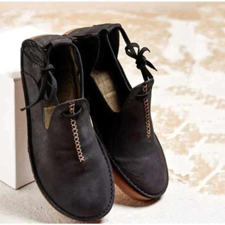 Chaussures d’automne pour femme en cuir suédé noir, avec bout arrondi, coutures contrastantes apparentes et lien décoratif sur le côté, présentées sur un fond clair pour un style moderne et élégant.
